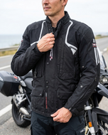 Rider fastening the collar of the black Helite Touring airbag jacket, standing beside an adventure motorcycle by the sea